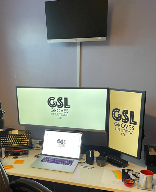 Photo of a desk with a MacBook Pro in the centre. There is a wide screen positioned above the MacBook screen, and a portrait format screen to its right. To the left of the MacBook is an external keyboard mounted near vertical and a red box with controls on the front beneath it. On the wall high above the wide monitor is another screen.